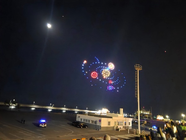Multiple colorful clusters of drones orbiting like tiny planets around a glowing center over the Santa Cruz port.