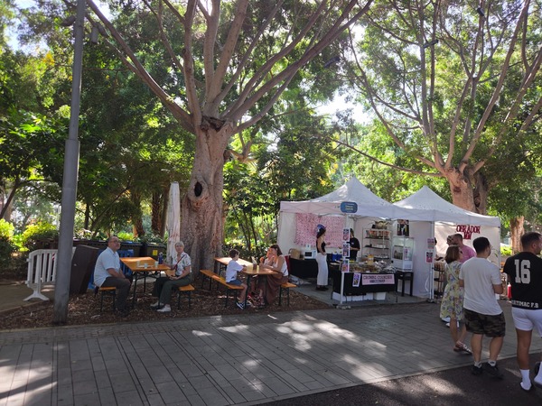 Dessert stalls with Crumble Cookies and shaded seating under big ficus trees. Taken at Plenilunio 2025 in García Sanabria Park, Santa Cruz de Tenerife.