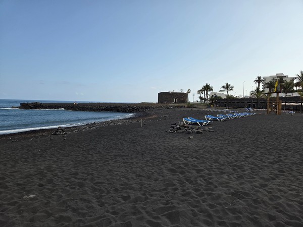 Wide view of Playa Jardín with black volcanic sand, calm waves and the small fortress in the distance.