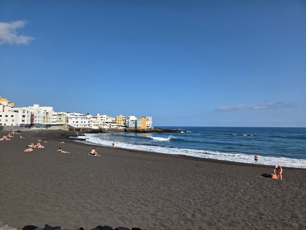 Sunbathers spread out on the spacious black beach of Punta Brava under clear skies.