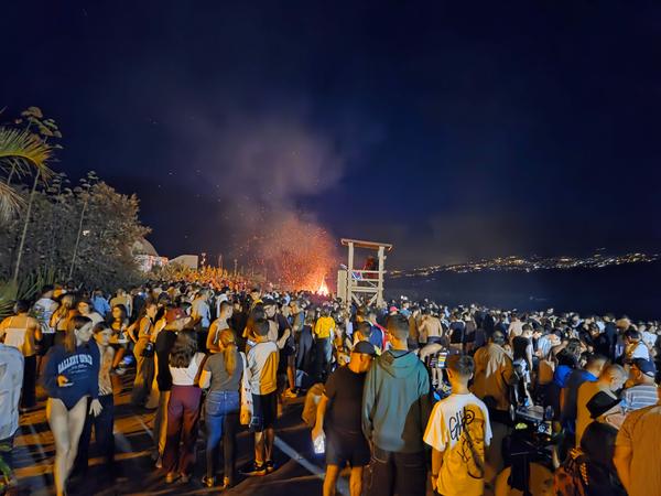 A large crowd facing a bonfire near the beach during the San Juan festivities with a night sky backdrop.