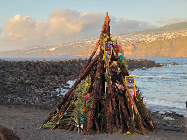 A decorated wooden bonfire structure on the beach at sunset, with a human-shaped figure and colorful ornaments for San Juan.