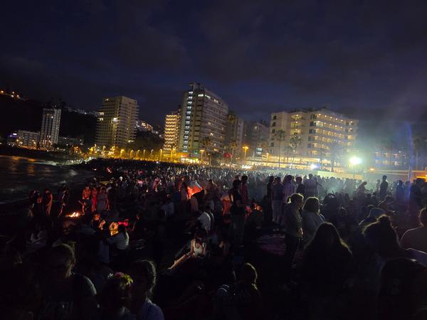 A dense crowd gathered on the beach at night, illuminated by firelight and city lights during the San Juan Festival.