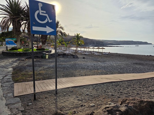 Accessible ramp and wooden walkway leading onto the black-sand beach at Playa San Juan, with a wheelchair sign and palms nearby.