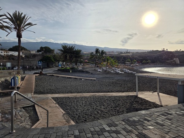 Sunbeds and straw parasols set on the black-sand shore of Playa San Juan with calm water.