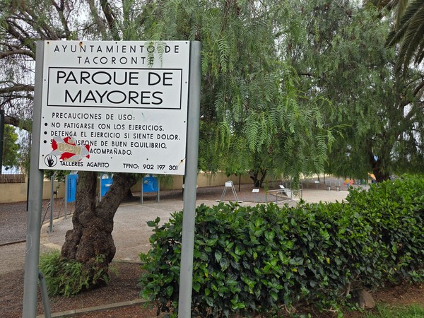 Sign for Parque de Mayores in Tacoronte with outdoor exercise equipment shaded by trees.