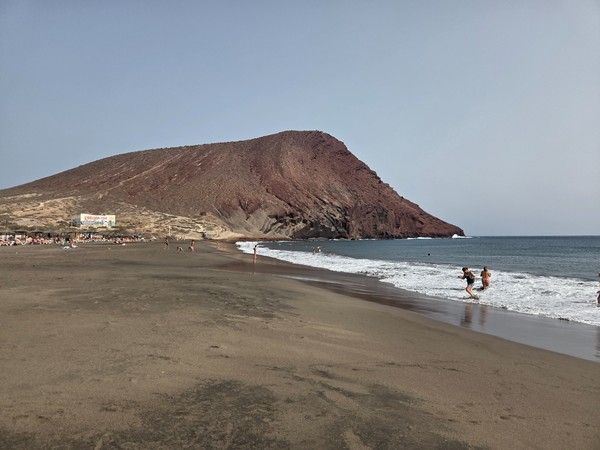 Close view of Montaña Roja rising above La Tejita’s shoreline, with bathers along the wet sand and small waves rolling in.