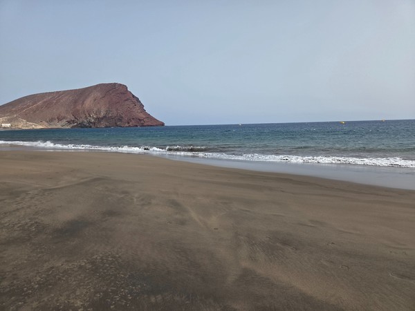 Aerial-style panorama of La Tejita Beach with long dark-sand shoreline and Montaña Roja beyond, showing lifeguard posts and sunbed rows.